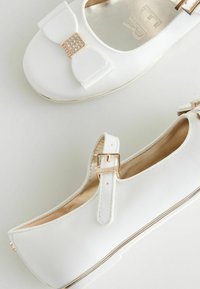 Ballerine bianche con finitura satinata, caratterizzate da un grande fiocco decorato con strass e una fascia metallica con fibbia dorata.