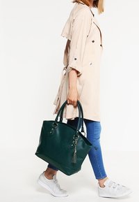Femme en trench-coat beige et jean bleu marchant, portant un grand sac à main vert foncé avec un détail de pompon.