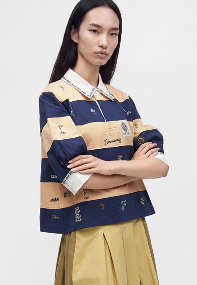 Femme aux longs cheveux foncés portant une chemise rayée bleu marine et beige avec des détails brodés et une jupe plissée jaune, bras croisés.