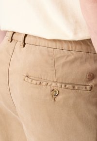 Pantalon en coton beige avec une poche arrière dotée d'une fermeture à bouton et d'un détail de couture, porté avec une chemise de couleur claire.