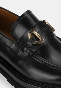 Zwarte leren loafers met een gladde textuur, goudkleurige gespdetails en contrasterende stiksels langs de onderrand.