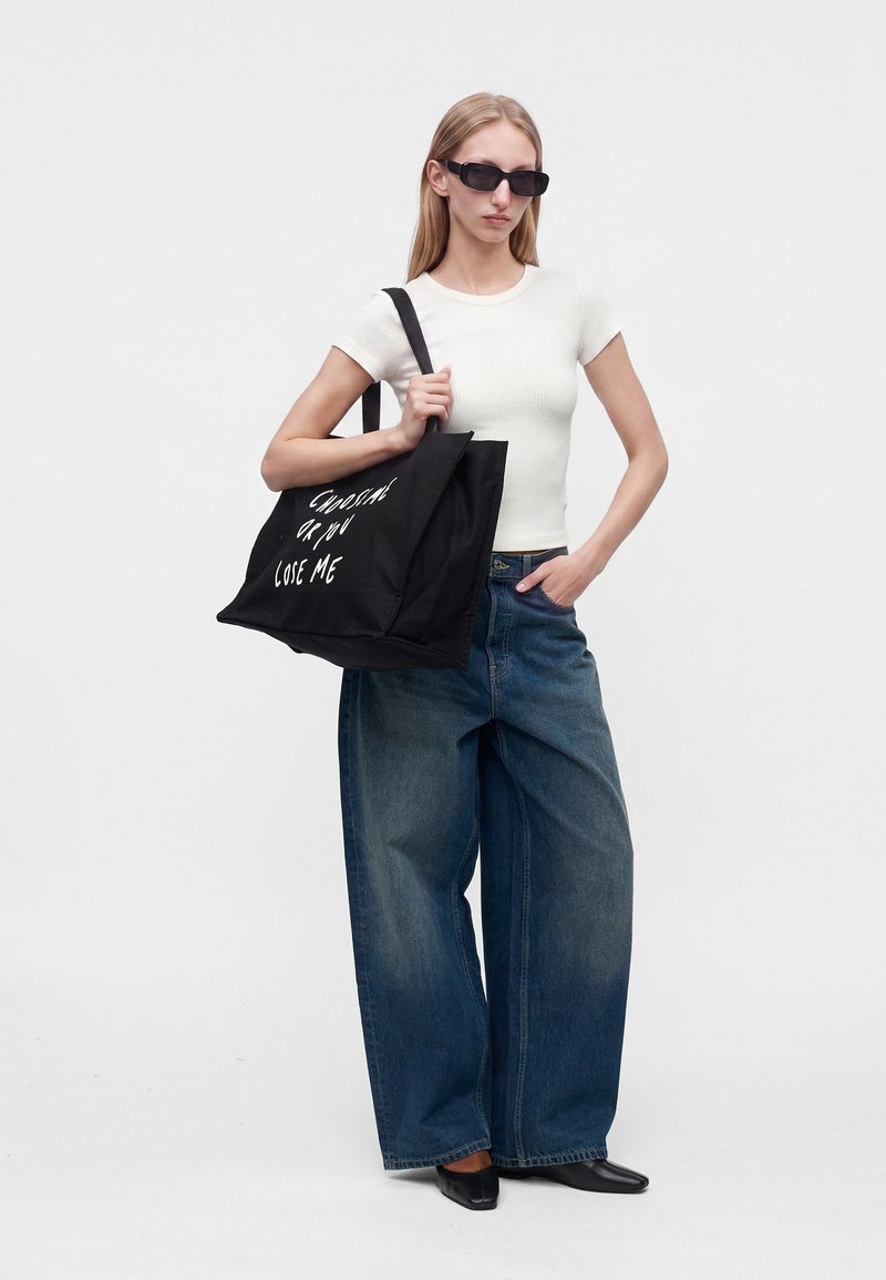 Jeune femme portant des lunettes de soleil, un haut blanc à manches courtes, un jean à jambes larges, des chaussures noires, tenant un sac noir avec un texte blanc.