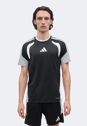 Tânăr cu păr închis și creț, purtând o ținută sportivă Adidas neagră și gri cu logo în față, în picioare pe un fundal simplu și deschis.