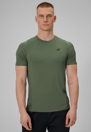 Hombre atlético con cabello corto que lleva una camiseta ajustada de manga corta 4F de color verde oliva y pantalones negros, de pie delante de un fondo gris liso.