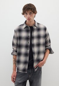 Camisa de franela a cuadros en blanco y negro con botones en la parte delantera, mangas largas y un solo bolsillo en el pecho, llevada sobre una camiseta negra.