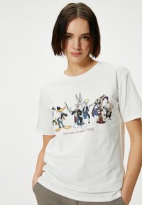 T-shirt en coton blanc arborant un graphique coloré de personnages de Warner Bros. avec le texte "100 ANS DE WARNER BROS." en bas.