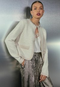 Hellbeige, kurz geschnittene Strickjacke mit weicher Textur, rundem Halsausschnitt, Perlenbesatz und aufgesetzter Tasche, kombiniert mit einem metallisch plissierten Rock.