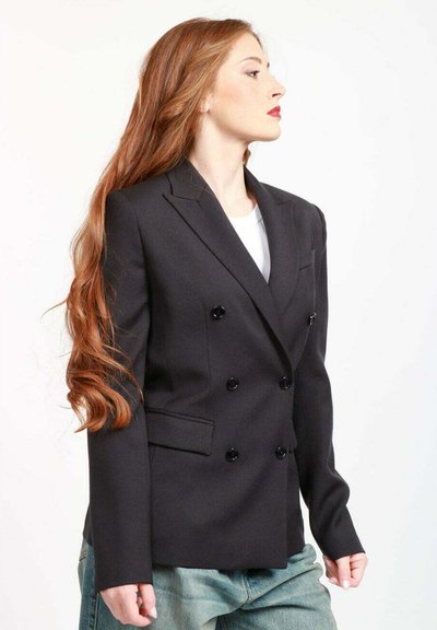 Blazer nero doppiopetto con rever a punta, dotato di due tasche frontali e bottoni neri. Indossato sopra una maglia bianca e pantaloni azzurri chiari.