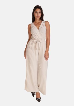 Beige wikkel-jumpsuit met een strikceintuur, verticale plooien, V-halslijn en brede pijpen. Gecombineerd met zwarte puntige hakken met strikdetails.