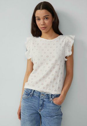 Top en coton blanc avec des manches courtes à volants, ornée de broderies en œillets et d'un col rond. Associé à un jean bleu clair.