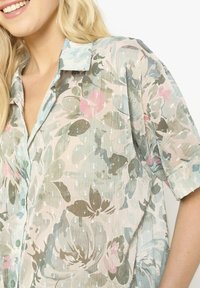 Woman wearing a pastel floral patterned button-up shirt with short sleeves and a collar, smiling, with long blonde hair.