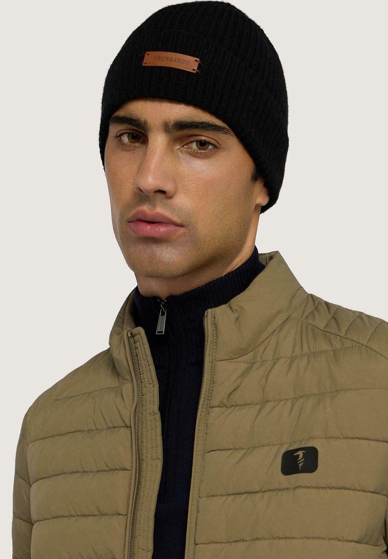 Cappello a coste nero in maglia con toppa in pelle. Giacca imbottita marrone chiaro con cerniera. Maglione blu scuro sotto. Design pulito e minimalista.