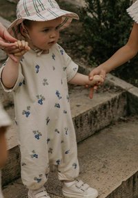 Lys beige romper med drueprint, struktureret stof, løs pasform, elastiske manchetter, parret med hvide sneakers. Barn som holder hænder med et andet.