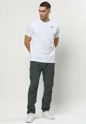 Mężczyzna w białej koszulce z krótkim rękawem z logo, ciemne spodnie typu cargo i białe trampki, stoi z rękami za plecami, patrząc w lewo.