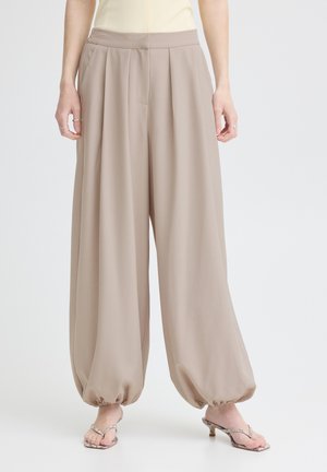 Frau trägt beige weite Hose mit gerafften elastischen Bündchen und Sandalen mit Schlangen-Print und Absatz, steht vor weißem Hintergrund.