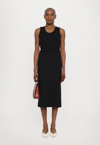 Schwarzes ärmelloses Oberteil mit Rüschendetails, kombiniert mit einem knielangen schwarzen Bleistiftrock, weißen spitzen Schuhen und einer kleinen braunen Handtasche.