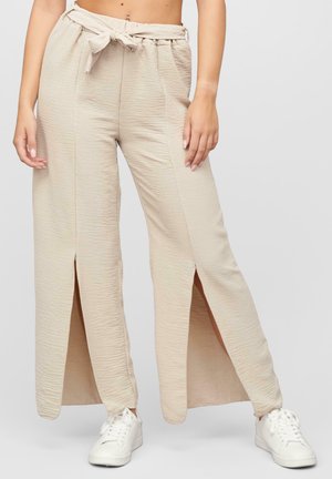 Pantalons larges beiges en tissu texturé, avec des fentes à l'avant et une taille à nouer. Associés à des baskets blanches.