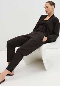 Woman wearing black pants, black tank top, and black bomber jacket sits relaxed in a modern white chair with hands in pockets.