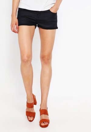 Frau trägt schwarze Shorts, steht mit einer Hand in der Tasche, trägt rostfarbene Sandalen mit Absatz vor weißem Hintergrund.