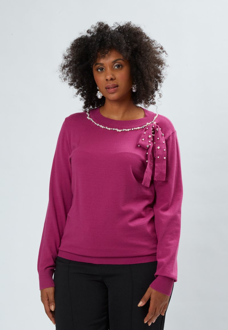 Maglione fucsia con scollo rotondo, caratterizzato da un bordo in perle e un accentuato fiocco sulla spalla sinistra. Maniche lunghe con polsini a coste.