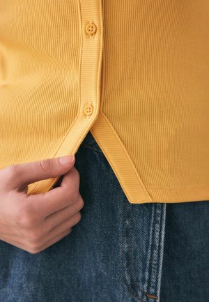 Close-up af en hånd, der justerer kanten på en gul ribstrikket skjorte med knapper over blå denimjeans.