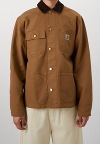 Giacca da lavoro marrone in cotone con colletto in velluto a coste, chiusura frontale con bottoni, due tasche sul petto e due tasche inferiori. Dotata di finiture in metallo color oro.