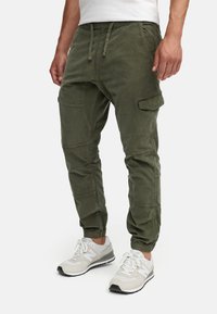 Pantaloni cargo verde oliva con polsini elasticizzati e vita con cordoncino. Presentano tasche laterali e una vestibilità rilassata. Abbinati a scarpe da ginnastica grigio chiaro.