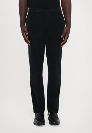 Uomo che indossa pantaloni di velluto a coste neri e mocassini in pelle nera, in piedi davanti a uno sfondo chiaro e uniforme.