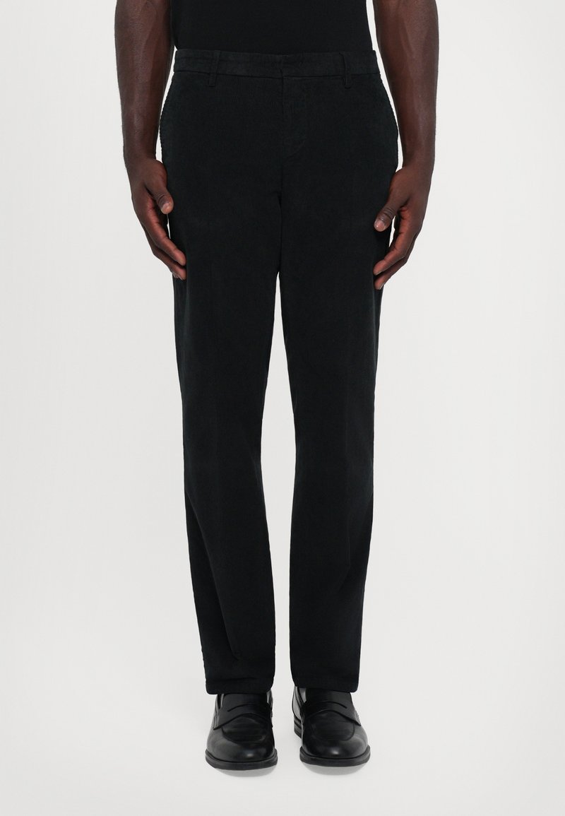 Hombre vestido con pantalones de pana negros y mocasines de cuero negros, de pie frente a un fondo claro y sencillo.