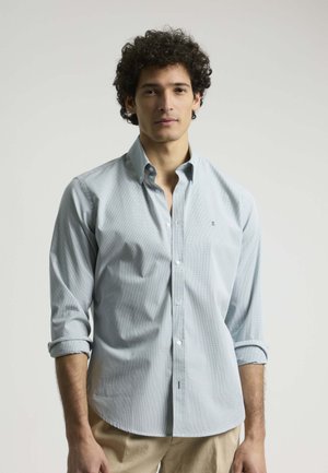 Camisa de botón azul claro con un pequeño patrón de cuadros, mangas largas enrolladas en los puños y un sutil logo en el pecho.
