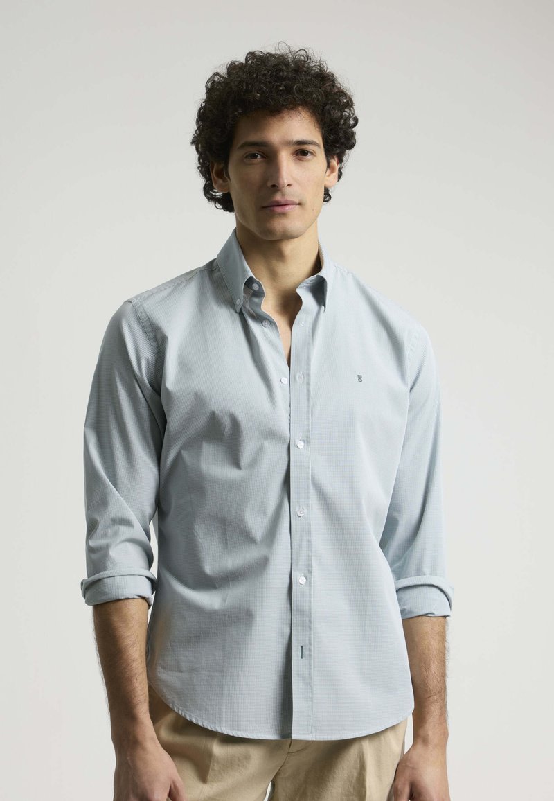 Camisa de botón azul claro con un pequeño patrón de cuadros, mangas largas enrolladas en los puños y un sutil logo en el pecho.