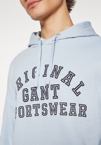 Gros plan d'une personne portant un sweat à capuche bleu clair avec l'inscription « ORIGINAL GANT SPORTSWEAR » en lettres noires et en gras sur la poitrine.
