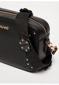 Bolso cruzado de cuero negro con textura de guijarro, cierre dorado, detalles de tachuelas, acentos en forma de flor y correa de tela ajustable con broche dorado.