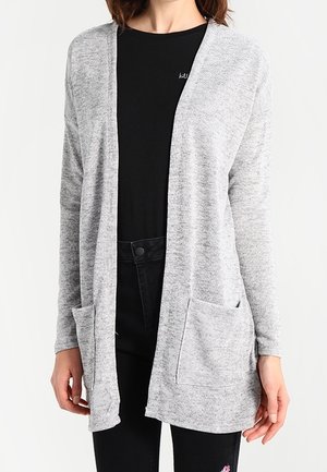 Lichtgrijze cardigan met open voorkant, lange mouwen en twee zakken aan de voorkant, gedragen over een zwart shirt en zwarte broek.