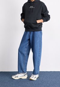 Svart huva sweatshirt med framficka och vitt logo, parat med ljusblå baggy denimjeans och flerfärgade sneakers.