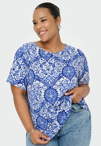 Leende kvinna iklädd en blommig blå och vit kortärmad topp, kombinerad med ljusblå jeans och silverörhängen.