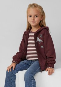 Burgunderfarbiges Zip-Hoodie mit Kapuze, verziert mit einem silbernen Pailletten-Pferdemotiv. Gestreiftes Hemd darunter und blaue Jeans. Weicher, leicht strukturierter Stoff.