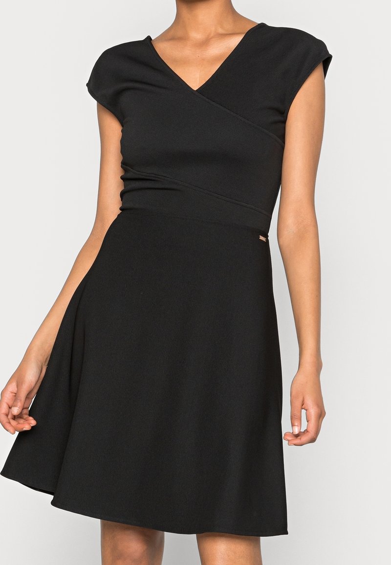 Femme portant une robe noire sans manches avec un corsage en V style portefeuille et une jupe fluide en ligne A, debout devant un fond clair uni.