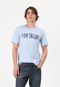 Gaiši zils kokvilnas T-krekls ar apaļu dekoltē un īsām piedurknēm, ar melnu grafisku apdruku, kurā redzams "TOM TAILOR" un cits teksts.
