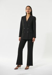 Blazer nero doppiopetto con bottoni prominenti, abbinato a pantaloni neri su misura. Tessuto liscio, vestibilità strutturata e pantaloni a campana.