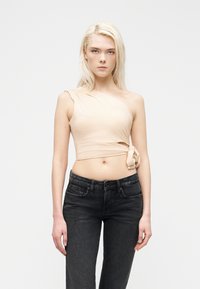 Ξανθιά γυναίκα που φοράει μπεζ cropped τοπ με έναν ώμο, δεμένο στη μέση, και μαύρα τζιν ψηλόμεσα, στέκεται μπροστά σε μια απλή φόντο.