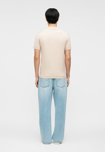 Homme debout, dos à l'appareil photo, portant un polo beige à manches courtes et un jean large bleu clair, sur un fond uni.