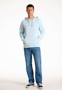 Sudadera con capucha de color azul claro con el texto "COPENHAGUE", bolsillo frontal, combinada con jeans de lavado claro y zapatillas negras. Textura suave.