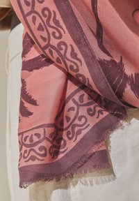 Écharpe rose et violette avec bordure à motifs et dessins de feuilles, posée sur un tissu blanc avec bord effiloché visible.