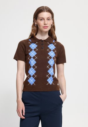 Kvinde står med den ene hånd i lommen, iført en brun kortærmet polosweater med blåt argylemønster og marineblå bukser.
