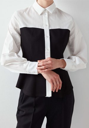 Personne portant une chemise noire et blanche à manches longues boutonnée avec un col blanc et un pantalon noir, ajustant un poignet avec l'autre main.