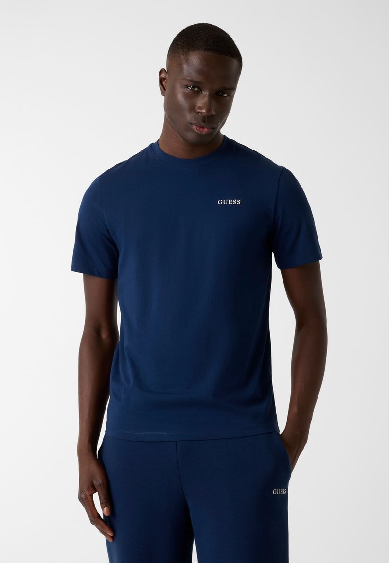 T-shirt en coton bleu marine avec un col rond et des manches courtes, arborant un petit logo blanc "GUESS" sur le haut de la poitrine à gauche.