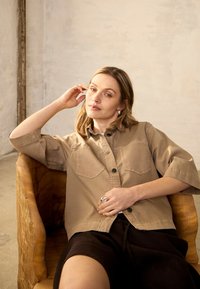Femme aux cheveux blonds portant une chemise boutonnée beige et un short noir, assise confortablement dans une chaise en bois contre un mur uni.