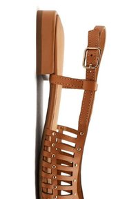 Sandalia de cuero marrón con tacón plano, que presenta un diseño de tiras, acentos de tachuelas metálicas y una correa para el tobillo ajustable.