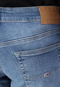 Ljust blå denimjeans med en läderlapp med texten "TOMMY JEANS." Har rundade bakfickor och kontrasterande sömmar. Texturen ser något använd ut.